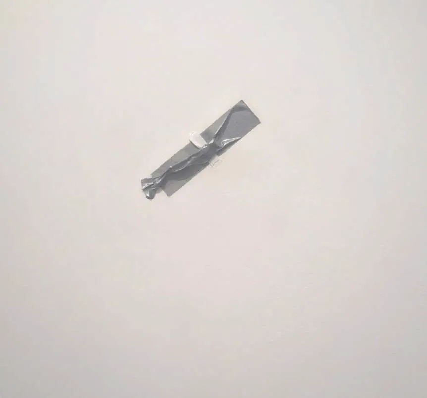 Beyaz, boş bir duvarda, yukarıdan aşağıya doğru hafifçe çapraz yerleştirilmiş gümüş rengi bantla tutturulmuş, Maurizio Cattelan'ın ünlü duvara bantlanan muz eserindeki muzun küçük, soluk renkli bir bölümü yakın çekim olarak görünüyor.