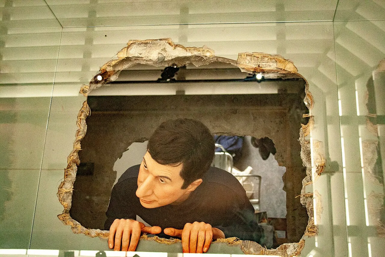 Maurizio Cattelan'ın tanınan eserlerinden biri olan, gri-beyaz bir duvar veya tavandaki yırtık, düzensiz bir delikten meraklı bir ifadeyle yukarı doğru bakan, elleriyle deliğin kenarına tutunmuş, koyu saçlı gerçekçi bir erkek figürü heykeli görülüyor.