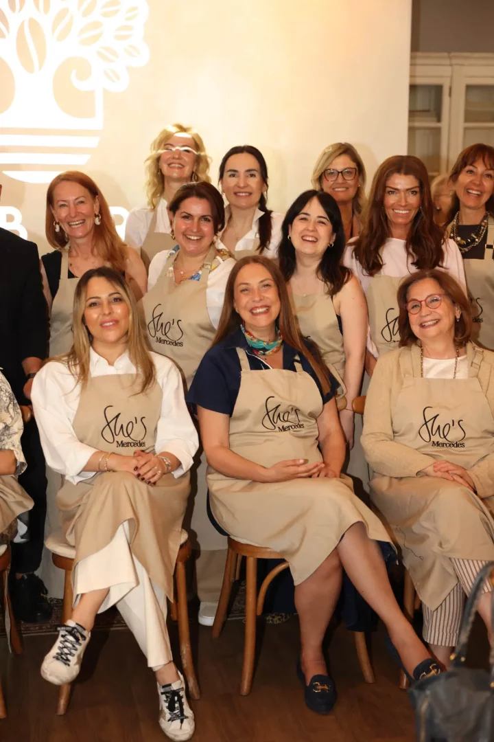Bir grup kadın, She's Mercedes logolu bej önlükler giymiş, gülümseyerek OgGusto etkinliğinde bir araya gelerek fotoğraf çekiliyor; bu neşeli an, markanın gastronomi dünyasına verdiği önemi ve kadınların katılımını vurguluyor.