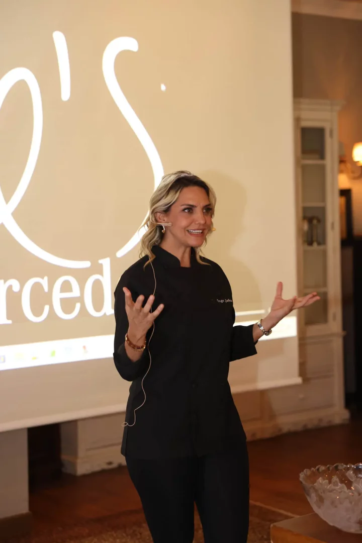 Mercedes-Oggusto gastronomi etkinliği sırasında sahneden konuşan, başındaki mikrofonu ve siyah şef ceketiyle gülümseyen, elleri açık bir şekilde anlatım yapan sarışın kadın, arka plandaki devasa logo yansıtması önünde bir sunum gerçekleştiriyor.