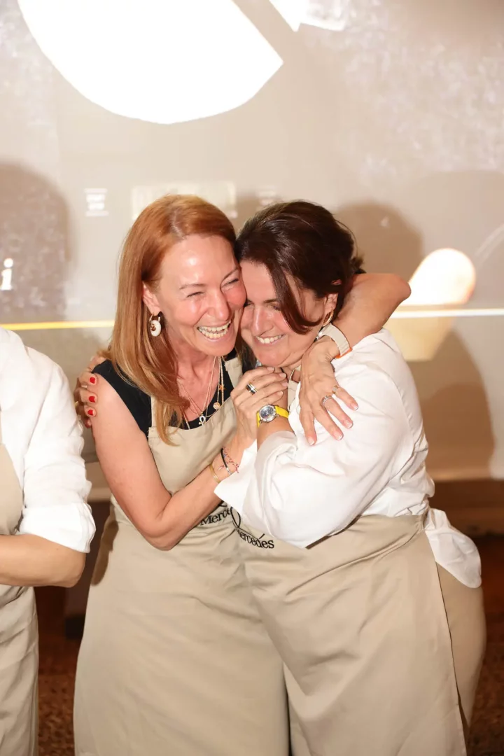 Mercedes-Oggusto gastronomi etkinliğinde, bej renkli Mercedes logolu önlükler giyen, biri kızıl diğeri koyu saçlı iki orta yaşlı kadın, birbirlerine sıkıca sarılarak ve tüm içtenlikleriyle kahkaha atarak etkinliğin coşkulu ve keyifli atmosferini yansıtan samimi bir anı paylaşıyor.