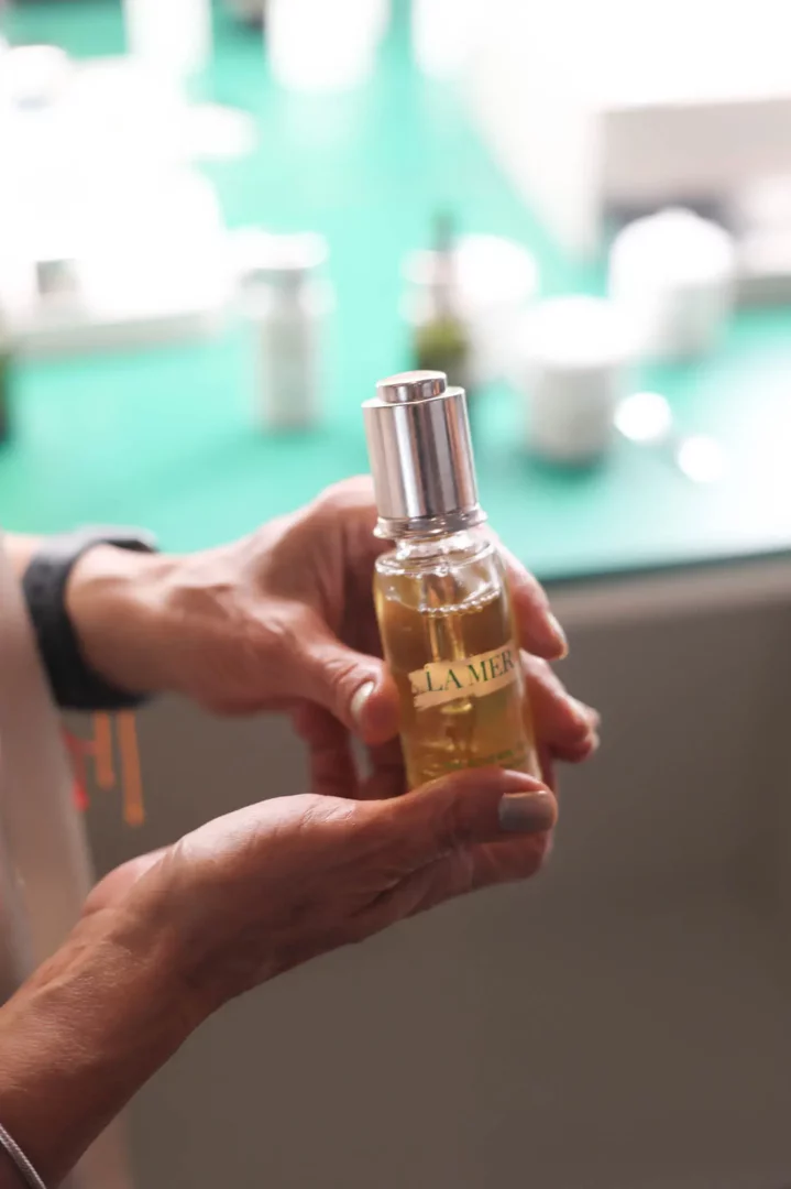 Yakından çekilmiş bu fotoğrafta, ojeli eller, üzerinde LA MER yazan, şeffaf camdan, altın sarısı serum içeren, gümüş pompalı bir kozmetik şişesini özenle tutuyor, lüks bir cilt bakımı etkinliği atmosferini yansıtıyor.
