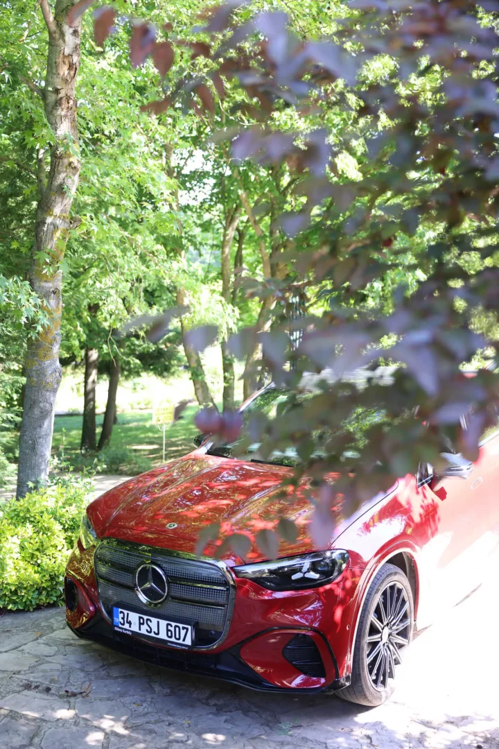 Doğal yeşillikler arasında, koyu kırmızı rengiyle dikkat çeken modern bir Mercedes-Benz otomobilin ön kısmı, test sürüşü rotasındaki bir molada ağaç yapraklarının arasından gün ışığında zarifçe yansırken görünüyor.
