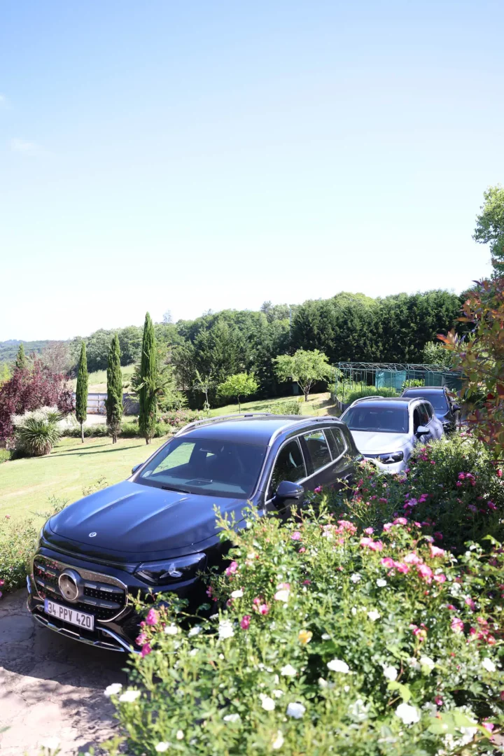Çiçek açan pembe ve beyaz güllerle dolu yeşil çalılıkların arasından görünen, 34 PPV 420 plakalı koyu renkli bir Mercedes-Benz SUV, arkasında sıralanmış iki gri SUV ile birlikte yemyeşil ağaçlarla çevrili ferah bir dış mekan sürüş noktasında konumlanıyor.