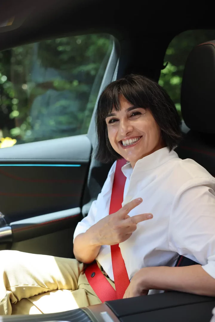 Mercedes aracında kırmızı emniyet kemeri takmış, neşeli bir gülümsemeyle poz veren, siyah saçlı bir kadın, elinde barış işareti yaparak sürüş deneyiminden keyif aldığını gösteriyor; arka planda yeşil doğa detayları görülüyor.