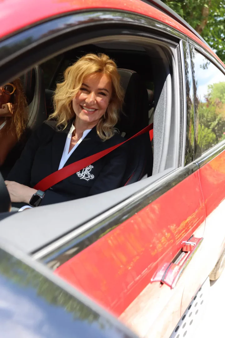 Mercedes-Benz’in She’s Mercedes buluşması kapsamında, kırmızı bir araçta oturmuş, üzerinde siyah blazer ve kırmızı emniyet kemeri olan sarışın, gülümseyen bir kadın güvenli sürüş deneyiminin keyfini çıkarırken kameraya gülümsüyor. Arka planda yeşil doğa detayları seçiliyor.