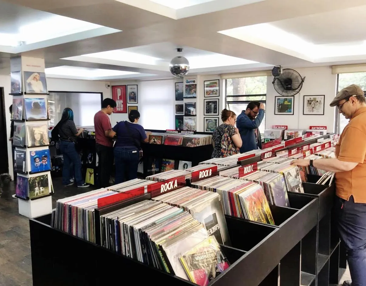 Canlı bir plak dükkanında, çoğu vinil plaklarla dolu uzun siyah raflar arasında müşteriler özenle albüm kapaklarını inceliyor; sağda şapkalı bir adam turuncu tişörtüyle plakları karıştırırken, arkada ise birkaç kişi daha geniş ürün yelpazesine göz atıyor.