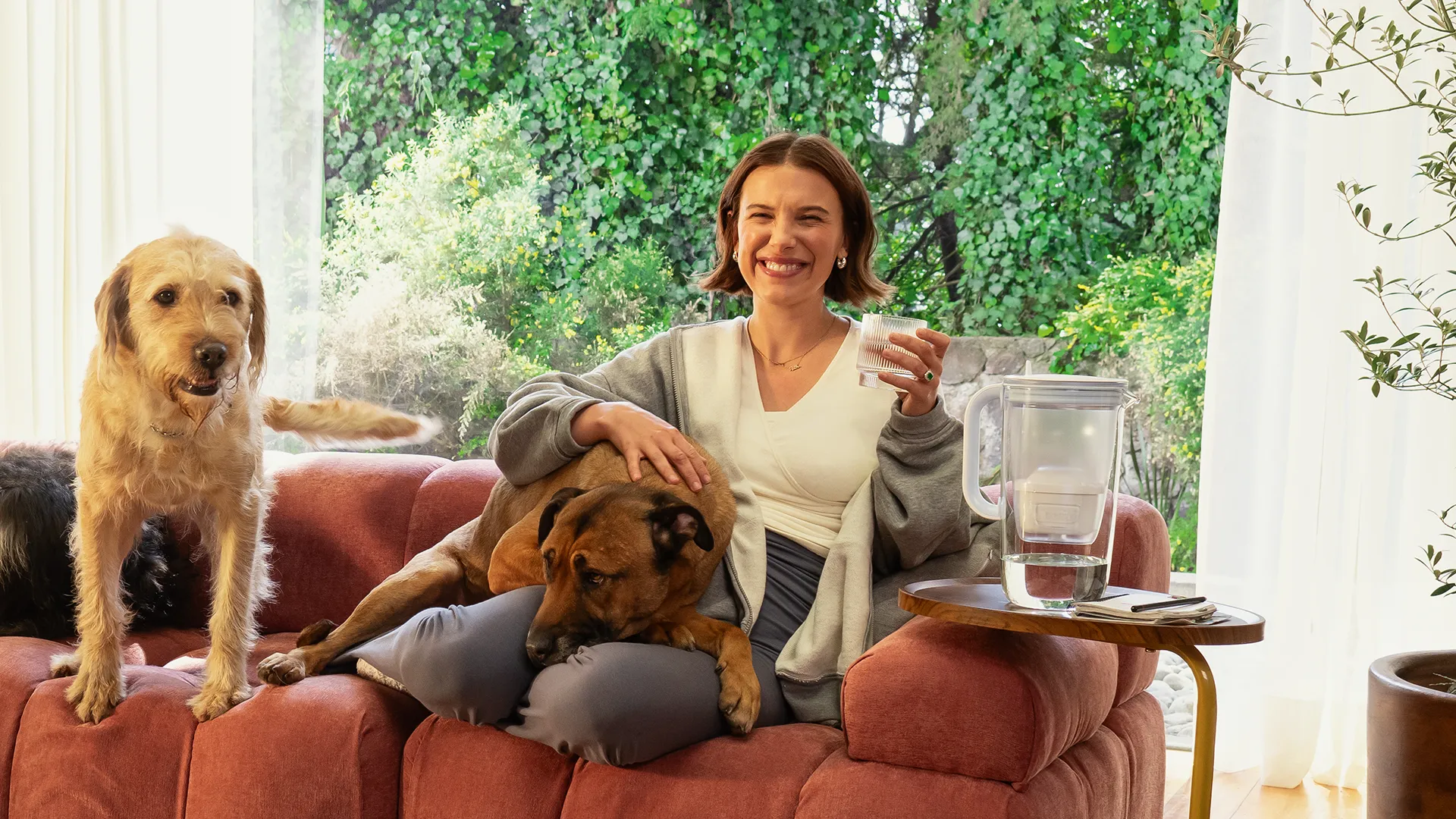 Kızıl kahve bir koltukta oturan Millie Bobby Brown, kameraya içtenlikle gülümseyerek elinde bir bardak su tutuyor; kucağındaki kahverengi köpeği okşarken, yanında duran tüylü açık renkli başka bir köpek de poz veriyor ve hemen sağındaki sehpada bir Brita su filtresi sürahisi bulunuyor, arka planda yemyeşil bitkilerle dolu bir pencere doğal bir atmosfer yaratıyor.