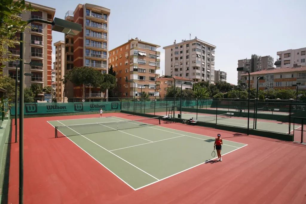 Miltas Spor Tesisleri'ne ait açık hava tenis kortlarından birinde, kırmızı ve yeşil zeminli sahada biri raket ve top tutan iki kadın oyuncu tenis oynamaya hazırlanırken, çevredeki yüksek katlı apartmanlar ve yeşillikler tesisin kentsel konumunu gözler önüne seriyor.