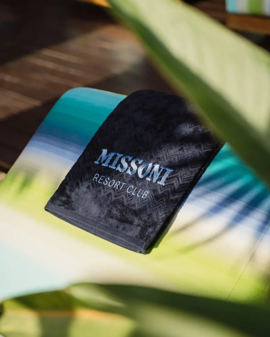 Güneşli bir ortamda, Missoni Resort Club yazılı, zarif zikzak desenli koyu renk bir havlu, Ibiza'daki Missoni Resort Club iş birliğini yansıtan mavi, turkuaz ve yeşil tonlarında çizgili bir şezlong minderinin üzerinde duruyor.