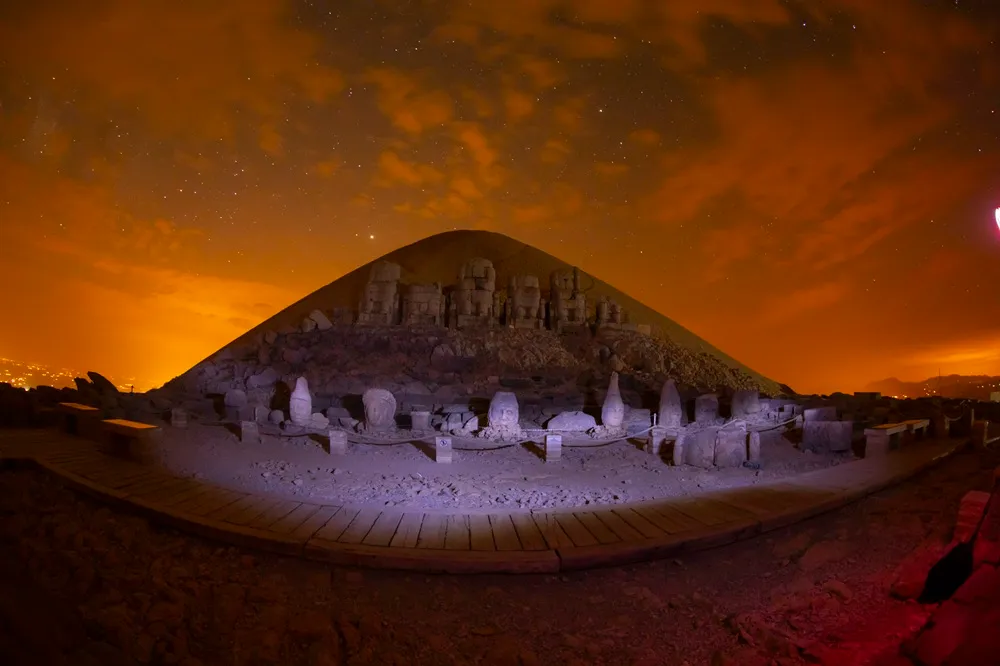 Nemrut Ören Yeri'ndeki devasa heykellerin ve yere düşmüş başların, yıldızlarla dolu kızıl turuncu bir gökyüzü altında, mor ışıklarla aydınlatılmış bir yürüyüş yolu önünden çekilmiş büyüleyici bir gece fotoğrafı.