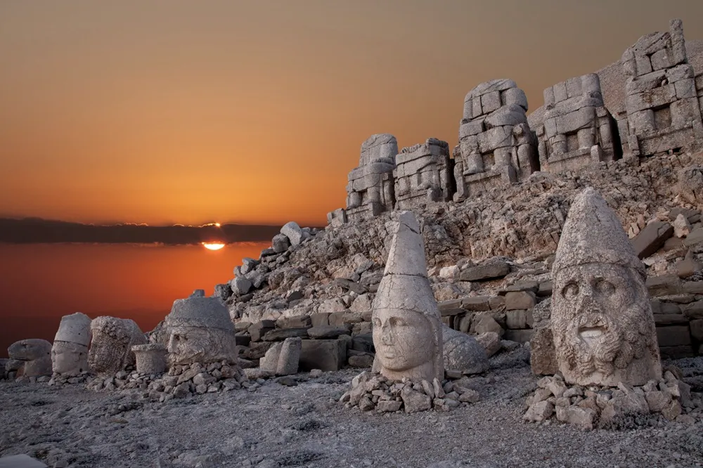 Kızıl ve turuncu tonlardaki gün batımında, bulutların arasından süzülen güneş ışınlarının aydınlattığı Nemrut Dağı ören yerinin kayalık yamaçlarında, Kommagene Krallığı'na ait devasa tanrı heykellerinin yıpranmış başları ve gövdeleri görünüyor.