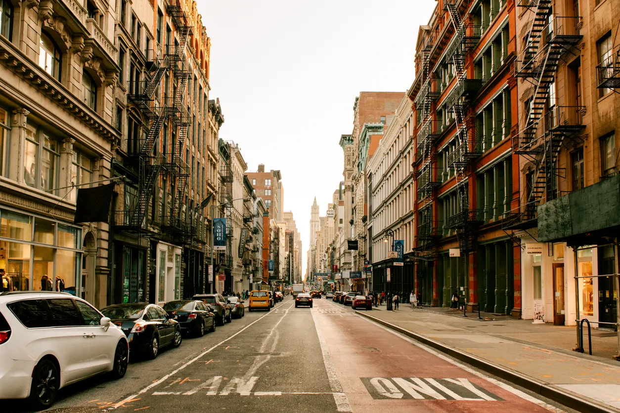 New York şehrinin hareketli bir caddesi, iki yanında dış yangın merdivenleriyle bezeli yüksek binaları, yol kenarında park etmiş çok sayıda arabası ve kaldırımlarda yürüyen insanlarıyla tipik bir kentsel manzarayı gözler önüne seriyor.