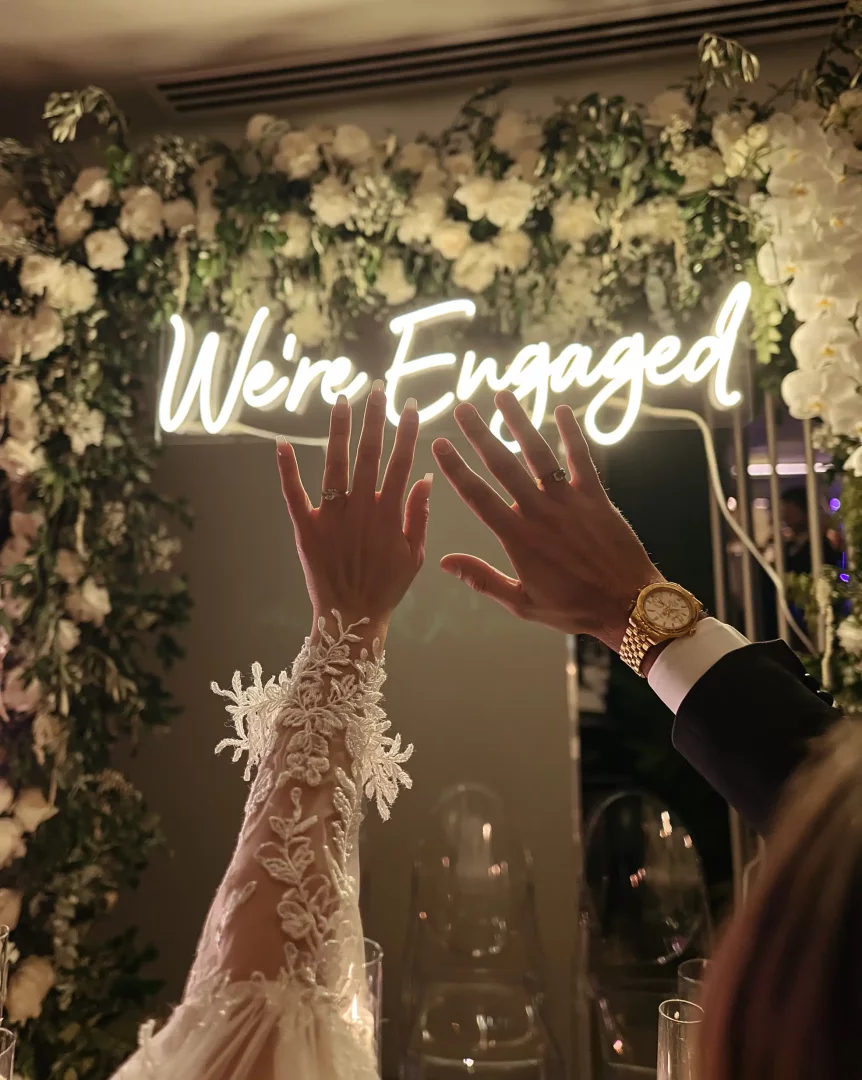 Nişan organizasyonu kapsamında özenle hazırlanan bir fonda, bembeyaz çiçeklerin ve We're Engaged yazan neon tabelanın önünde, yüzükleri takılmış ellerini coşkuyla kaldıran yeni nişanlı çift, mutlu bir anı kutluyor.