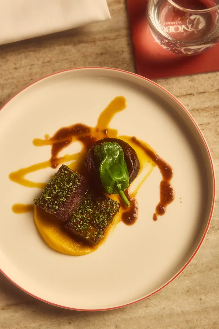 Nobu Matsuhisa'nın dünya çapında tanınan mutfak felsefesini ve Japon teknikleriyle Peru esintilerini harmanlayan yaklaşımını yansıtan bu özel sunumda, tabağın merkezinde otlar ve biberlerle kaplı iki parça Şili levreği, altında ipeksi sarı bir püre, yanında közlenmiş mor bir soğan ve üzerinde parlak yeşil bir biber ile zengin soslar, Nobu İstanbul'un sofistike gastronomik deneyimini vurguluyor.