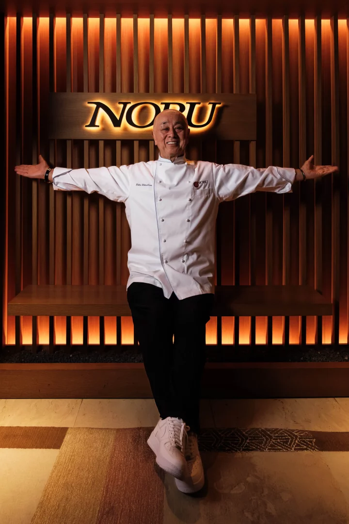 Şef Nobu Matsuhisa, beyaz şef üniforması ve spor ayakkabılarıyla, arkasında aydınlatılmış NOBU yazısının bulunduğu ahşap duvarın önünde kollarını iki yana açmış, Japon mutfağını bir yaşam stili haline getiren küresel gastronomi yolculuğunu yansıtan samimi bir gülümsemeyle poz veriyor.