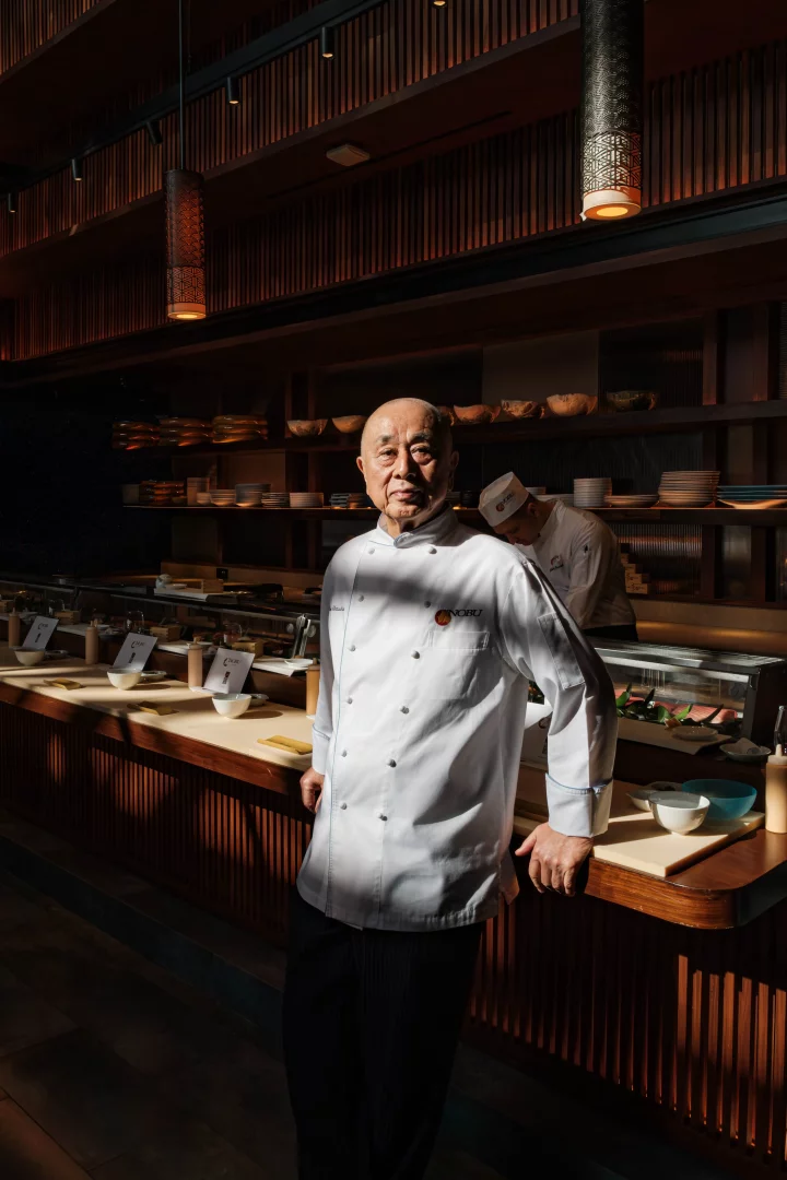 Dünyaca ünlü Şef Nobu Matsuhisa, sıcak ahşap paneller ve zarif aydınlatmalarla donatılmış modern Nobu restoranının mutfak veya suşi hazırlık alanında, kendine özgü mutfak felsefesini ve Japon mutfağına getirdiği bakış açısını vurgulayan bir kararlılıkla beyaz şef ceketi içinde duruyor.