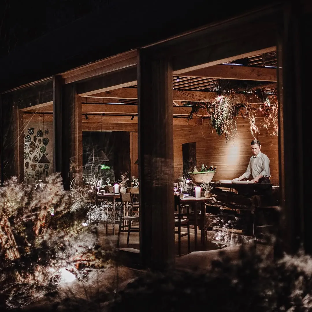 Noma restoranının iç mekanından bir gece görüntüsü; loş ışıklar altında, ahşap kirişlerle çevrili, masaların mumlar ve doğal bitkilerle süslendiği, tavandan sarkan kuru dalların ve sağda tezgaha eğilmiş bir görevlinin bulunduğu bu Kopenhag mekanının rustik ve zarif atmosferini dışarıdan gösteriyor.