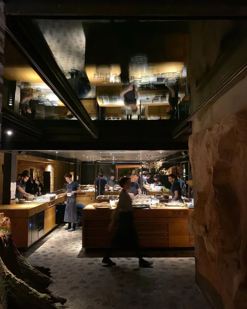 Noma restoranının loş ışıklı mutfağında, ahşap tezgahlar etrafında yemek hazırlayan Kopenhag ekibinin hareketli anları, tavanın yansıtıcı yüzeyinde tersine dönmüş bir şekilde yansırken, ön planda doğal ağaç kütüğü ve taş duvar modern ve rustik unsurları birleştiriyor.
