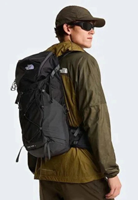Koyu renk şapka, güneş gözlüğü ve zeytin yeşili bir ceket giymiş, sırtında ip detayları ve The North Face logosu bulunan siyah bir doğa yürüyüşü sırt çantası taşıyan ve omzunun üzerinden hafifçe arkaya bakan bir adam, yeni bir macera için hazırlıklı görünüyor.