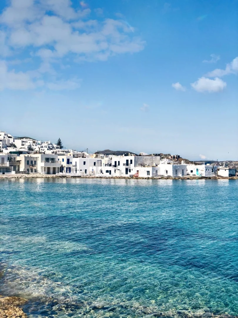 Güneşli bir günde, Yunanistan'ın Paros adasındaki pitoresk Naoussa köyünün masmavi ve berrak sularının önünde, tipik Kiklad mimarisine sahip, bembeyaz badanalı evler kıyı boyunca sıralanıyor.