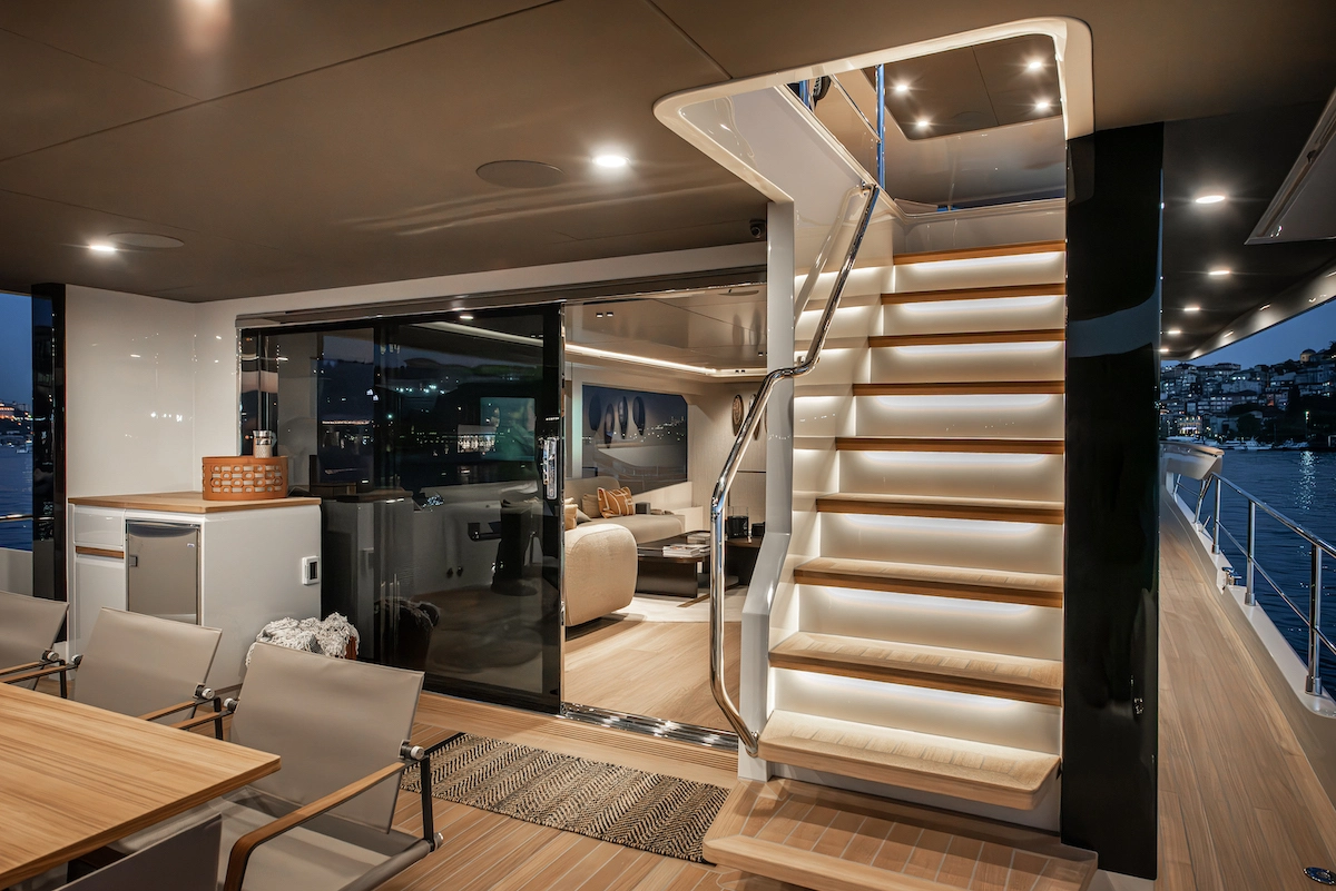 Numarine 27XP yatının sancak tarafındaki, basamak altı aydınlatmaları ve krom korkuluklarıyla dikkat çeken modern ahşap flybridge merdiveni, arkasında cam kapıdan görünen şık iç yaşam alanı ve uzakta parıldayan şehir ışıklarıyla akşam manzarasını gözler önüne seriyor.