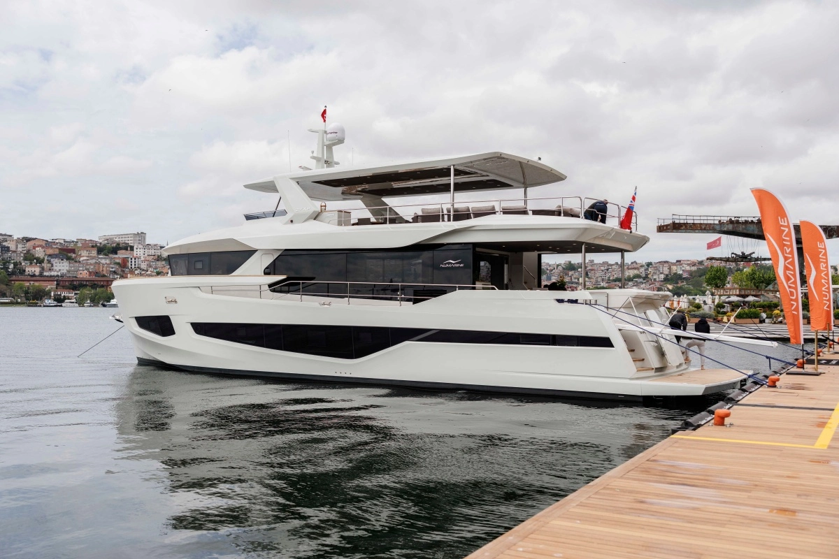 Gri bulutlu gökyüzünün altında, ahşap bir iskeleye bağlı duran şık beyaz Numarine 27XP explorer yat, koyu renk camları ve modern hatlarıyla dış tasarımının estetiğini ön plana çıkarırken, arka planda suyun karşısındaki tepelerde sıralanmış yerleşim yerlerinin siluetleri seçiliyor.