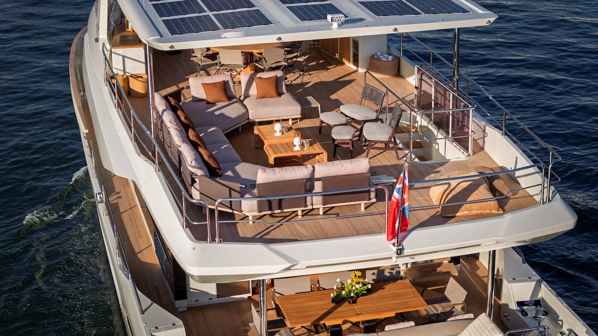 Numarine 27XP Explorer yatının flybridge'inin drone çekiminde, konforlu L şeklindeki koltuklar, dinlenme alanları, şık sehpa, ahşap zeminler ve çatıda güneş panelleriyle donatılmış geniş güvertesi berrak mavi denizde ilerlerken görülüyor.