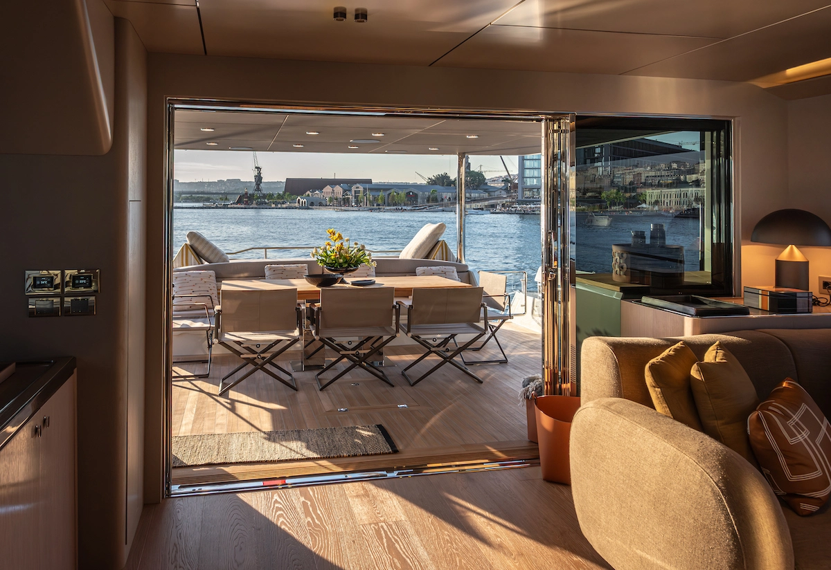 Numarine 27XP Explorer yatın modern salonundan bakıldığında, açık sürgülü kapının ardında, üzerinde sarı çiçeklerin olduğu yemek masası ve katlanır sandalyelerle donatılmış geniş arka havuzluk, uzaklardaki binalar ve vinçlerle dolu şehir silüeti ve sakin deniz manzarası ile birleşerek huzurlu bir yaşam alanı sunuyor.