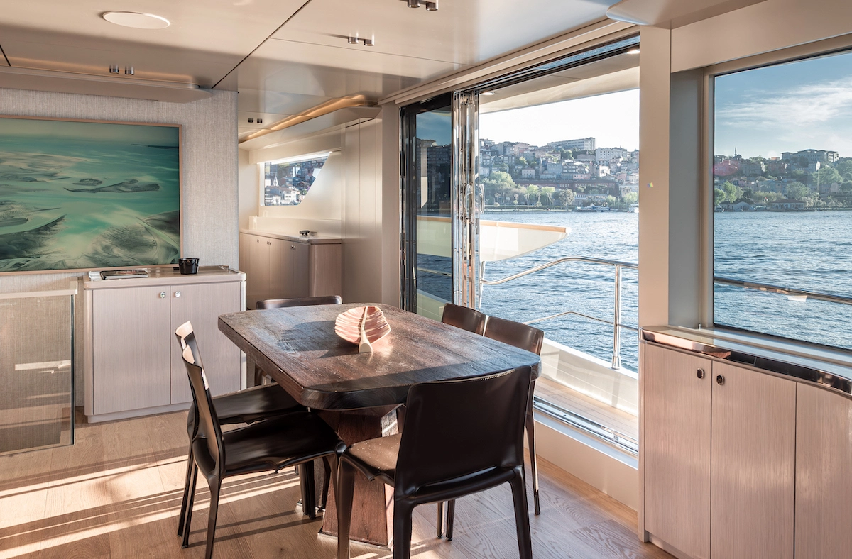 Numarine 27XP Explorer yatın geniş salonunda, güneş ışıklarıyla aydınlanmış ahşap yemek masası ve koyu renkli sandalyeler, dışarıdaki masmavi denizin ve karşı kıyıdaki şehrin panoramik manzarasını sunan geniş cam kapının önünde yer alıyor.