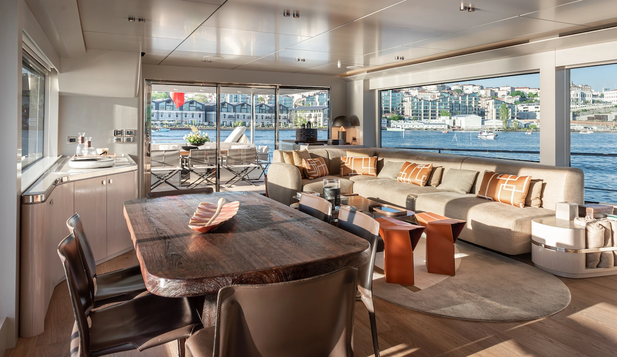 Numarine 27XP Explorer yatının lüks salonunda, ahşap yemek masası ve konforlu koltuk takımının bulunduğu geniş pencereden, güneşin aydınlattığı denizin ve karşı kıyıdaki şehrin muhteşem manzarası görülüyor.