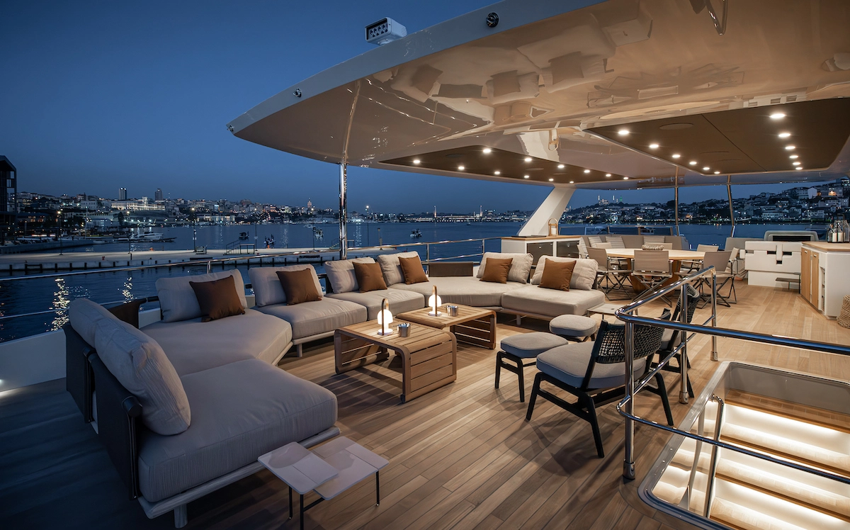 Numarine 27XP flybridge güvertesinde akşam ışıkları altında, rahat krem renkli koltuk takımlarının ve şık ahşap sehpalarda duran aydınlatmaların oluşturduğu modern oturma grubu ile geniş yemek masası ve arkadaki mini bar alanı, şehir manzarasına karşı lüks bir atmosfer sunuyor.