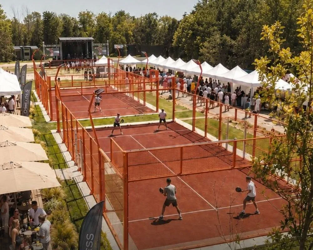 O'Padel Summer Fest etkinliğinde açık havada üç padel kortunda oyuncular maç yaparken, arkada müzik sahnesi ve çok sayıda beyaz çadırın etrafında toplanmış kalabalıklar festivalin canlı atmosferini yansıtıyor.