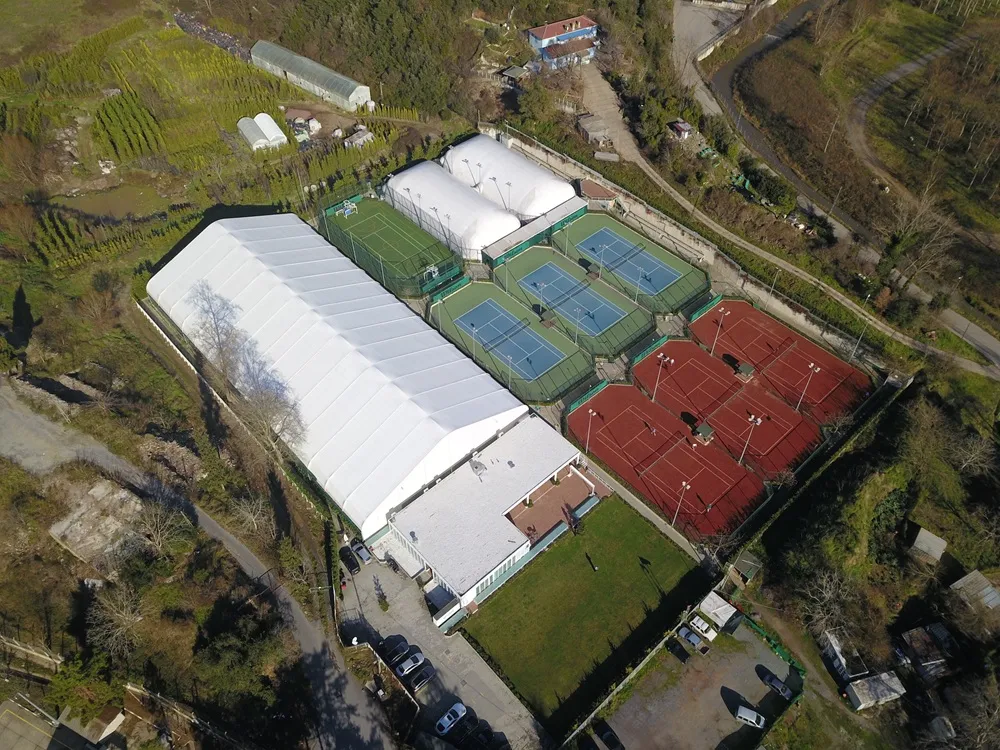 Optimum Tenis Akademisi'nin kuşbakışı bu geniş görüntüsünde, kırmızı toprak zeminli açık kortlar, mavi-yeşil akrilik açık kortlar ile büyük beyaz çadır ve kubbeli yapılar altında bulunan kapalı tenis kortlarının da yer aldığı kapsamlı bir spor tesisi, etrafı ağaçlarla çevrili yeşil bir alanda konumlanmış olarak görülüyor.