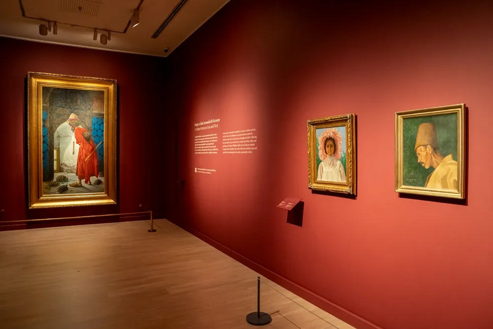 Koyu bordo renkli bir sergi salonu duvarında, solda büyük, altın varaklı çerçevede Osman Hamdi Bey'in ünlü Kaplumbağa Terbiyecisi tablosu, sağında ise sergiyi anlatan bir bilgilendirme paneli ve iki farklı portre tablosu dikkat çekiyor.