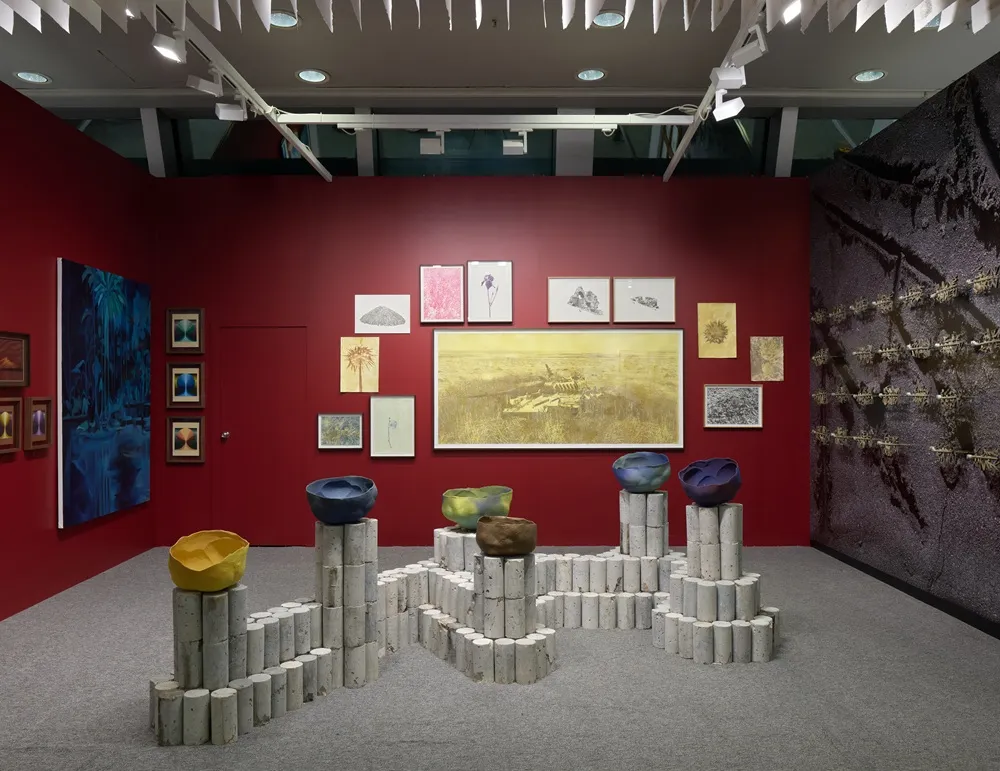 Otmar Uras'ın sanat eserlerinin sergilendiği kırmızı duvarlı ve gri halılı bir stantta, solda derin mavi bir orman tablosu, ortada büyük, sarı tonlarında bir peyzaj ve sağda koyu renkli dokulu bir duvar enstalasyonu çevrelerken, önde beton silindir kaideler üzerinde sarı, yeşil, mavi ve mor renkli, organik şekilli kaseler dikkat çekici bir düzenlemeyle sunuluyor.