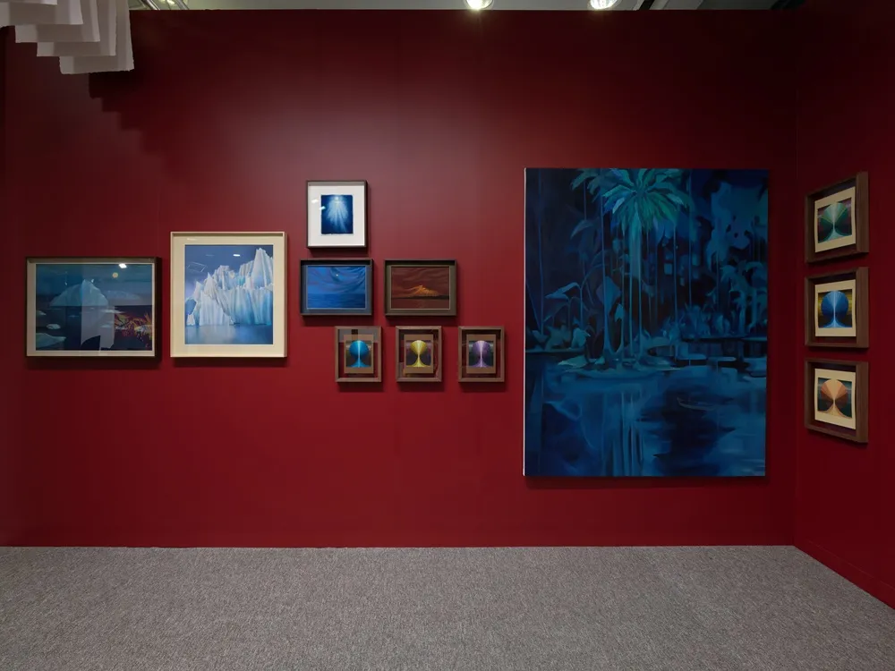 Koyu bordo renkli bir duvar üzerinde Otmar Uras'ın farklı boyutlarda eserlerinin sergilendiği bir sanat galerisi görünümü; solda buzdağları, ortada mavi tonlarda tropikal bir manzara ve sağda simetrik soyut desenler içeren çeşitli tablolar yer alıyor.