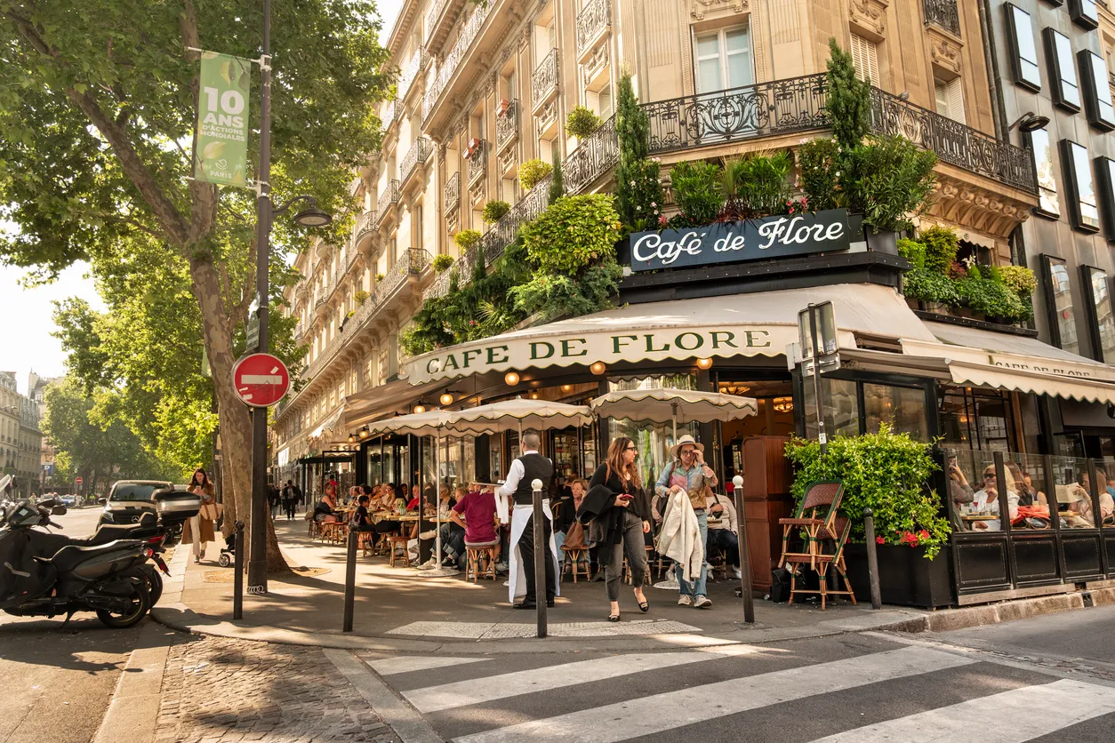 Paris'in ikonik Cafe de Flore'si, güneşe karşı uzanan tenteli ve şemsiyeli dış mekanında çok sayıda misafirin oturduğu, hareketli bir sokak köşesinde yer alıyor; klasik Paris mimarisinin süslediği üst katları ve kaldırımda park etmiş motosikletlerle canlı şehir atmosferini yansıtıyor.