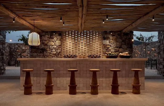 Paros'taki otantik bir bar mekanının akşam ambiyansını yansıtan bu geniş açılı görüntüde, taş örgü duvarlar ve doğal ahşap tavan kirişlerinin altında modern bir dokunuşla tasarlanmış, önünde altı adet konik ahşap taburenin sıralandığı zarif bir bar tezgahı görülüyor.