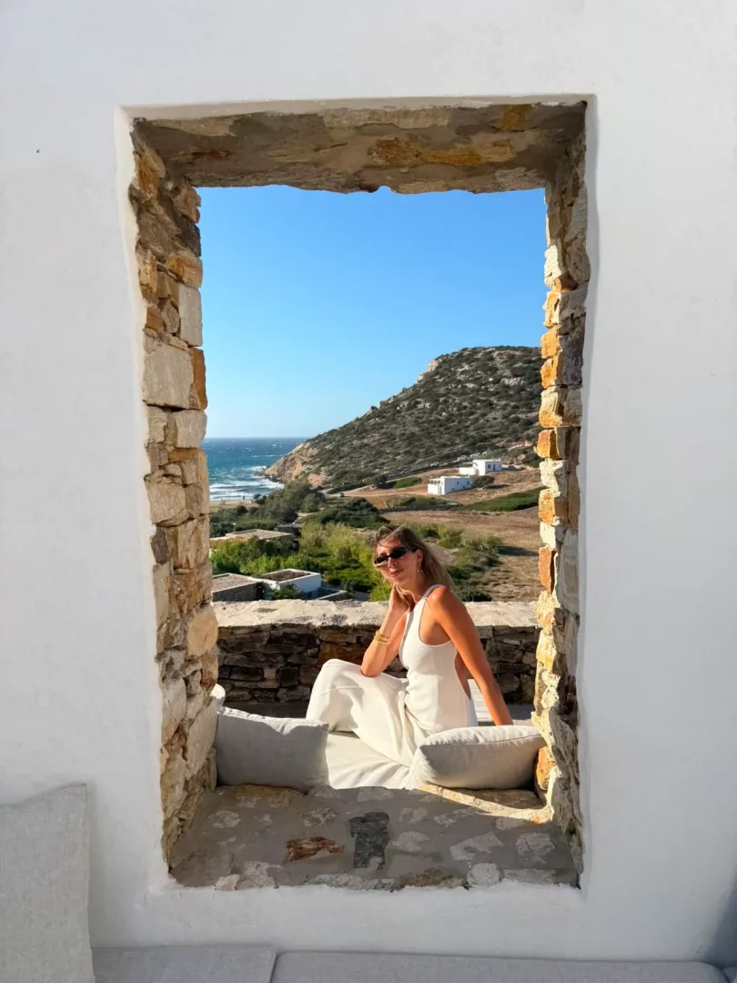 Paros'un karakteristik beyaz badanalı duvarları ve rustik taş pencere çerçevesi içinde, beyaz tulumu ve güneş gözlükleriyle oturan Cansın Denizer, arkasında uzanan masmavi Ege Denizi, dalgalı kıyı şeridi ve adanın yeşil tepeleriyle güneşli manzarayı keyifle izliyor.