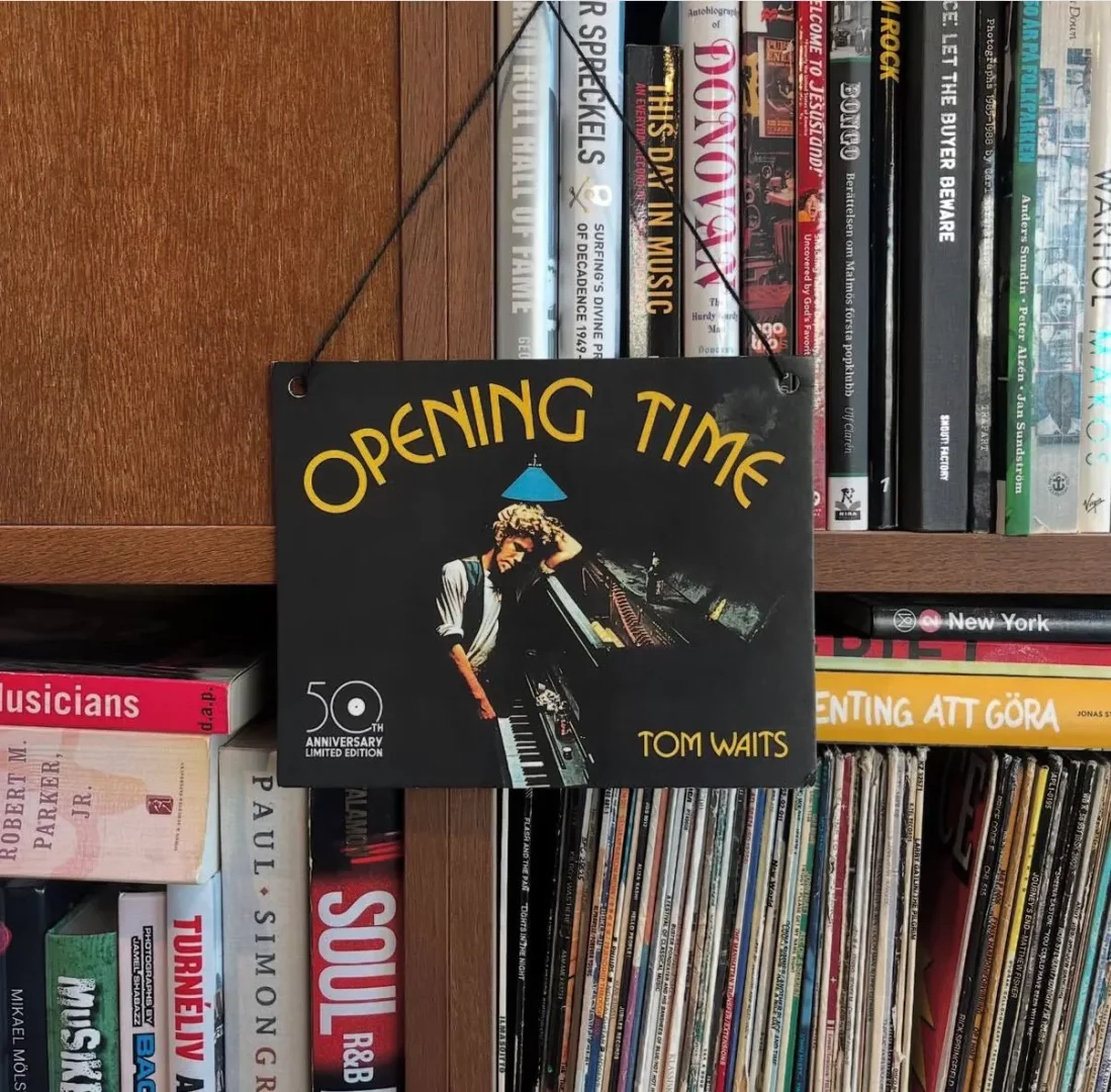 Siyah bir ip ile ahşap bir rafın önünde asılı duran, üzerinde Tom Waits'in piyanoya yaslanmış bir illüstrasyonu ve başında mavi bir abajur ile Opening Time yazısı bulunan, 50. yıl dönümü sınırlı üretim plaketi, arkasında müzik ve sanat kitaplarıyla dolu bir kitaplık ile eski plakların bulunduğu bir rafı gösteriyor.