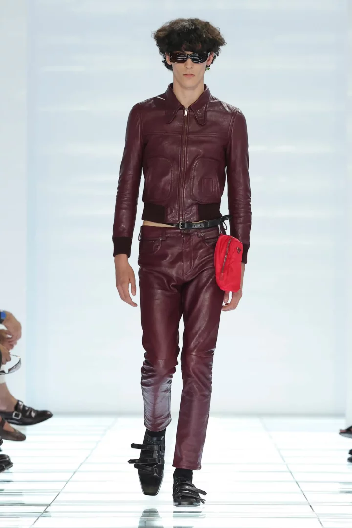 Prada İlkbahar-Yaz 2027 erkek defilesinde podyumda yürüyen model, bol cepli bordo deri bomber ceket, dar kesim deri pantolon, belinde kırmızı bir kemer çantası, futuristik güneş gözlükleri ve çoklu kayış detaylı siyah ayakkabılarıyla sade ama keskin bir erkek gardırobunu sergiliyor.