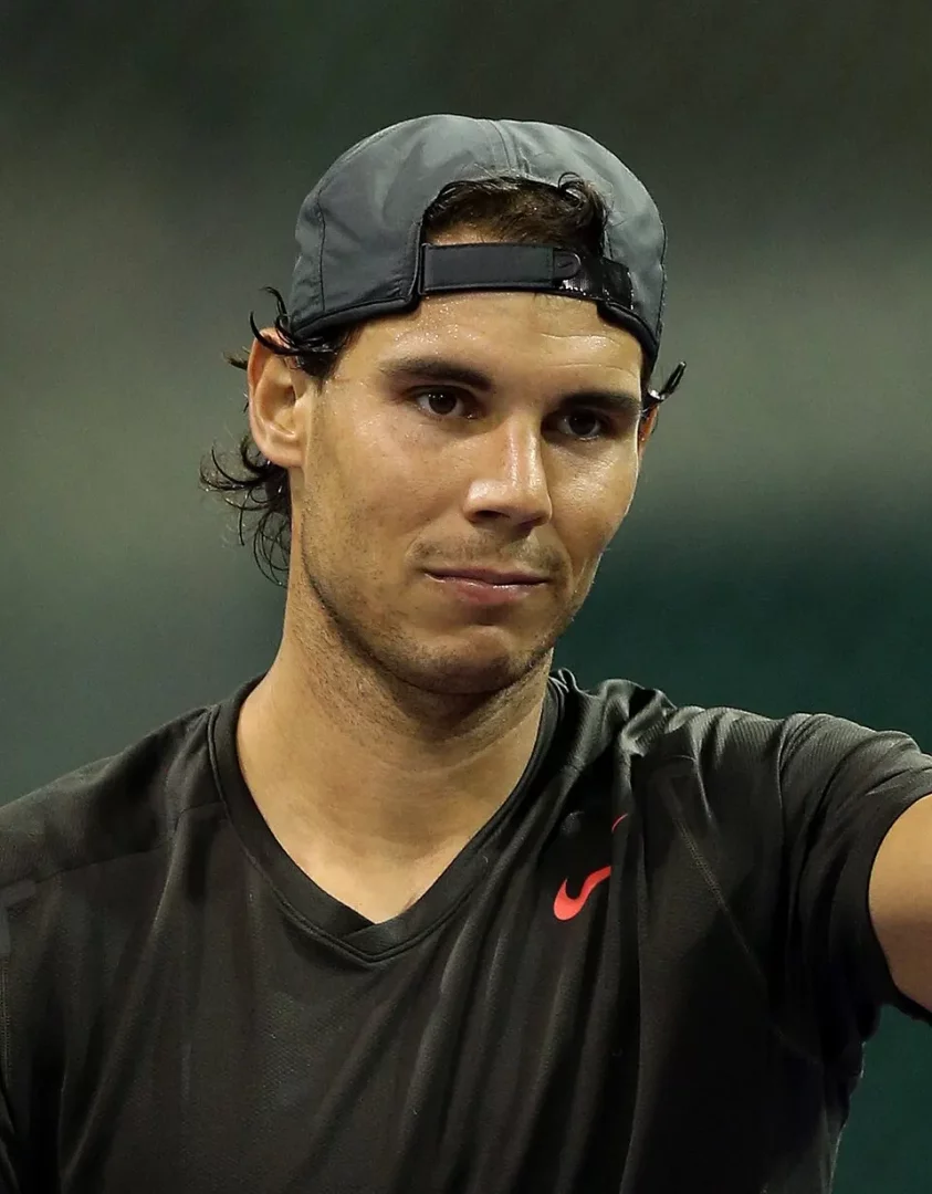 Şapkasını arkaya doğru takmış olan ünlü İspanyol tenisçi Rafael Nadal, siyah spor tişörtü ve belirgin Nike logosuyla, bir maç sonrası veya antrenman sırasında yüzündeki ter damlaları ve kararlı bakışlarıyla kameraya bakarken görüntüleniyor.