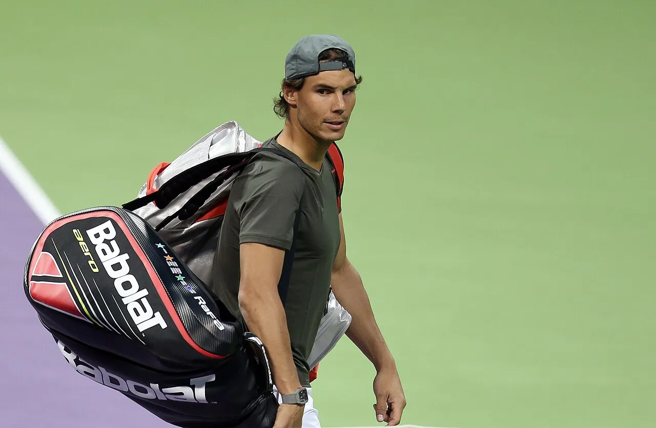 Ünlü tenisçi Rafael Nadal, gri şapkası ters takılmış ve koyu yeşil tişörtüyle, üzerinde Babolat logosu ve 'aero' yazısı bulunan büyük siyah-kırmızı tenis çantasını sol omzunda taşıyarak açık yeşil tenis kortunda ciddi bir ifadeyle yürürken doğrudan kameraya bakıyor.