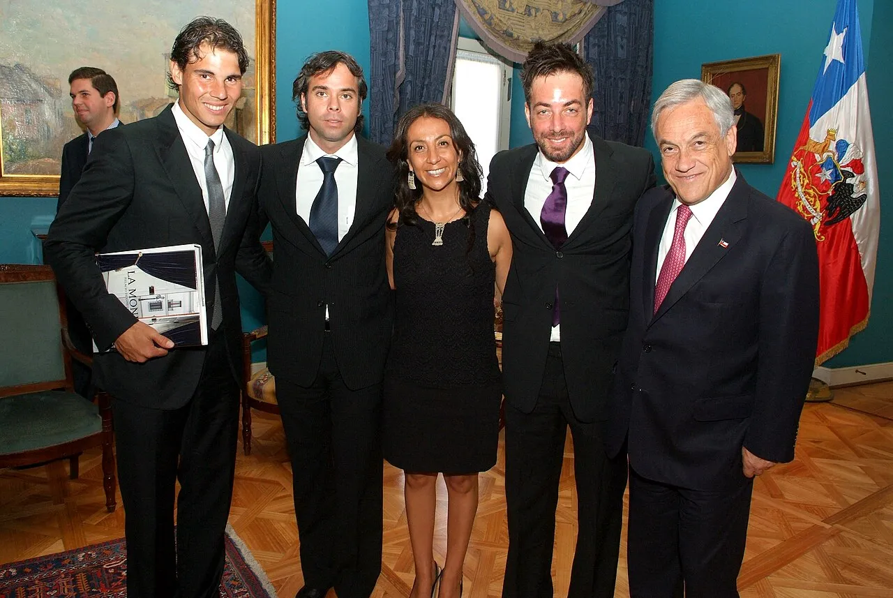 Rafael Nadal, koyu takım elbise ve gri kravatıyla solda duruyor, elinde açık renk bir kitap tutarken gülümsüyor; yanında üçü erkek biri kadın olmak üzere dört yetişkin daha var, en sağdaki beyaz saçlı adamın arkasında Şili bayrağı ve resmi bir resim çerçevesi görünüyor. Bu resim, Nadal'ın uluslararası temaslarını veya özel bir etkinliği yansıtan diplomatik bir toplantıda çekilmiş izlenimi veriyor.