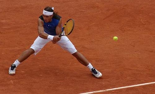 Rafael Nadal, toprak kortta geniş bir duruşla raketini savurmaya hazırlanırken, odaklanmış ifadesiyle beyaz kafa bandı, kol bantları ve şortuyla mavi atletini giymiş, tenis topu ise havada tam vuruş anını bekliyor.
