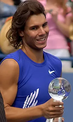 Ünlü İspanyol tenisçi Rafael Nadal, açık mavi Nike spor tişörtüyle, uzun dalgalı saçları ve içten gülümsemesiyle zaferini kutlarken, sağ elinde parıldayan şeffaf, küresel bir tenis kupasını gururla tutuyor.