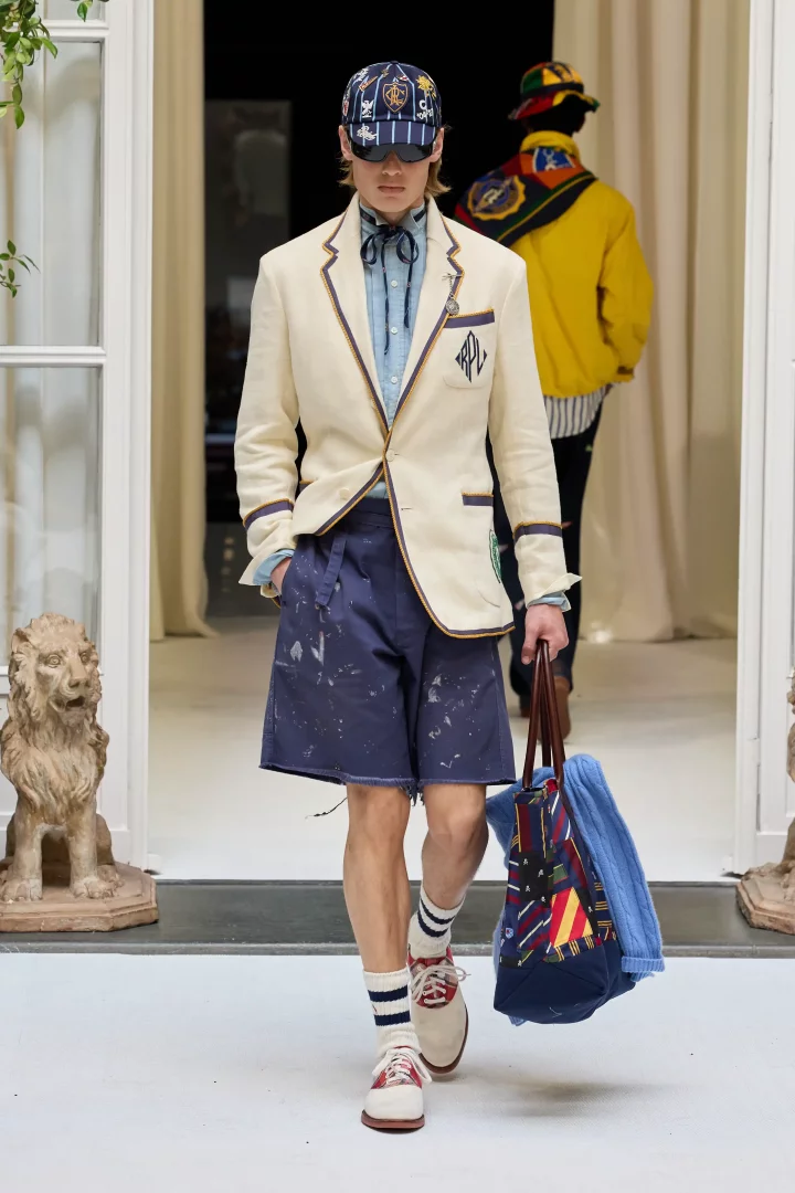Ralph Lauren 2027 İlkbahar-Yaz erkek giyim defilesinde, krem rengi kenarları lacivert ve altın detaylı blazer ceket, lacivert desenli şapka, açık mavi gömlek, beyaz boya sıçramalı lacivert şort ve çok renkli çantasıyla podyumda yürüyen model, Amerikan kampüs kültürü ve yeni Amerikan zarafetini yansıtan sportif ve rahat bir İlkbahar-Yaz 2027 erkek giyim görünümünü sergiliyor.