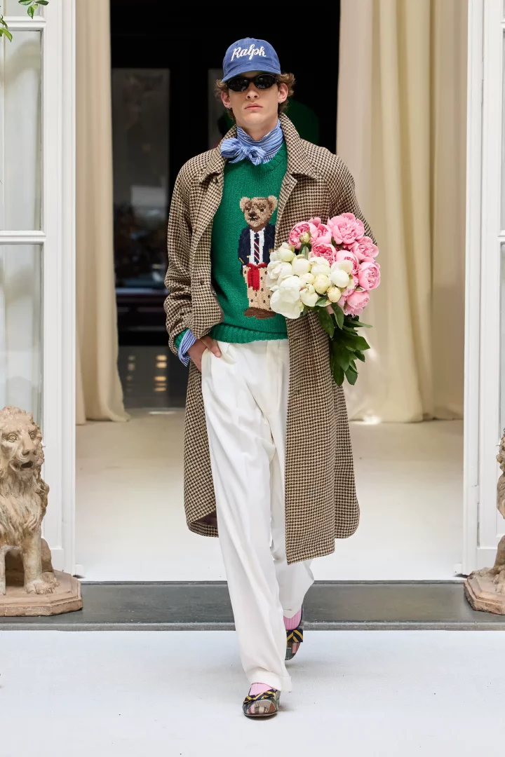 Ralph Lauren'in İlkbahar-Yaz 2027 erkek giyim koleksiyonundan bir görünüm sergileyen, mavi şapkalı, güneş gözlüklü bir erkek model, ekose uzun bir palto, üzerinde Polo Bear figürlü yeşil bir kazak ve bol beyaz pantolon giyerek, pembe ve beyaz şakayık buketini taşırken podyumda yürüyor.