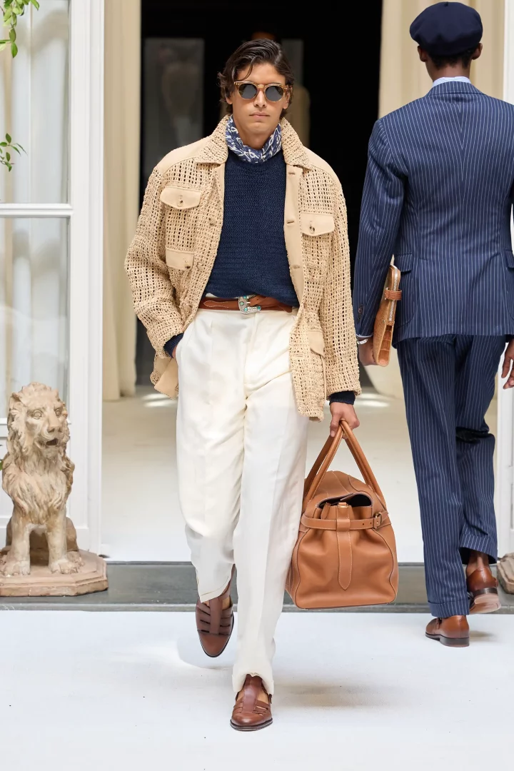 Ralph Lauren İlkbahar-Yaz 2027 erkek giyim koleksiyonundan, hafif terzilik ve Akdeniz estetiğini vurgulayan, örgü bej ceket, lacivert kazak, beyaz bol paça pantolon ve kahverengi sandalet giymiş, güneş gözlüklü bir model, elinde kahverengi deri çanta ile podyumda rahat ve şık bir duruşla yürüyor.